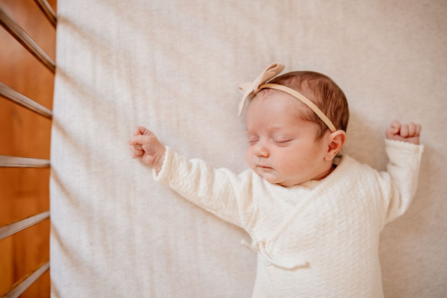 A sleeping baby wearing a white sweater lies peacefully on a cream-colored blanket with arms stretched outward.