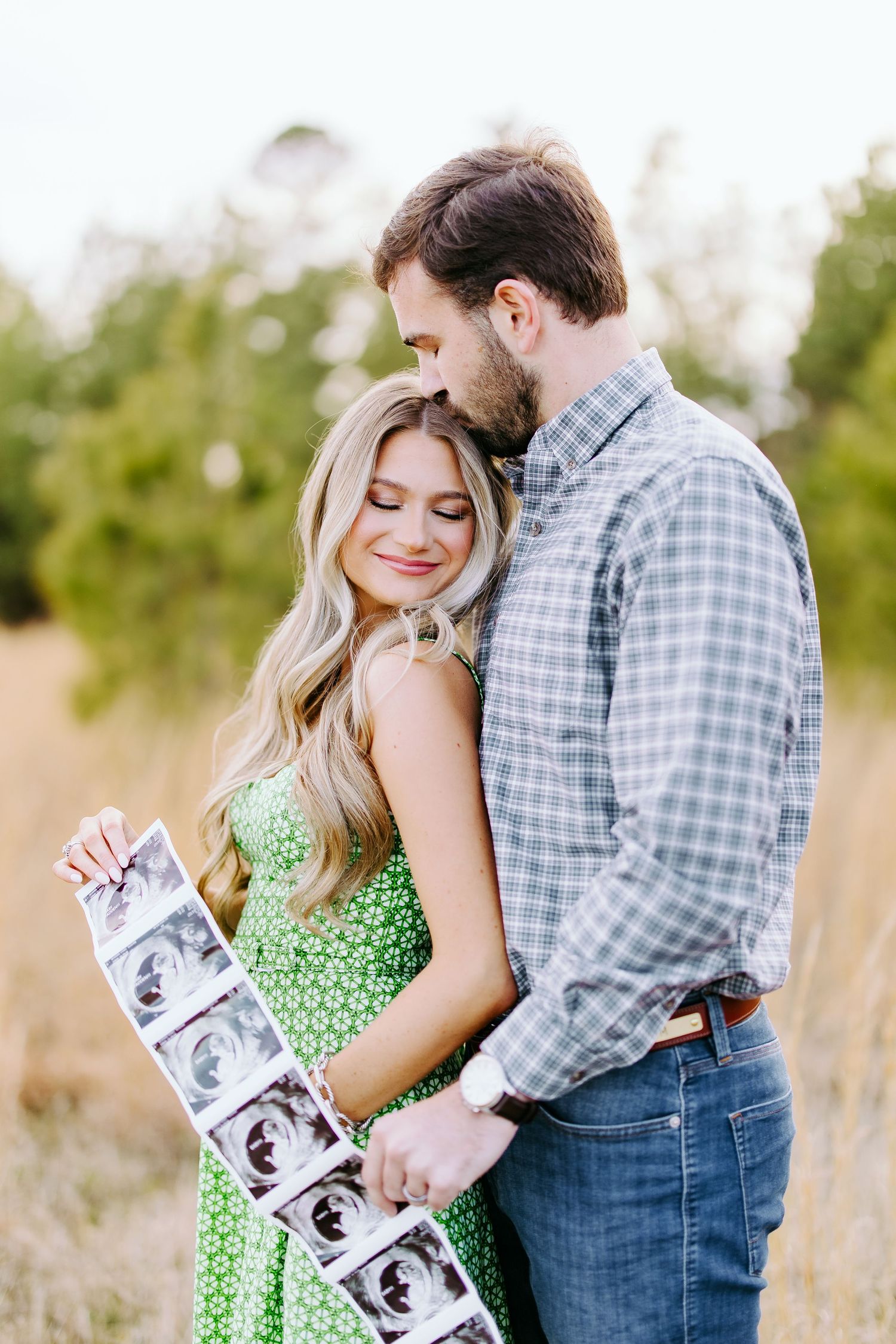 A couple stands together outdoors holding ultrasound photos in casual attire during a pregnancy announcement photoshoot.