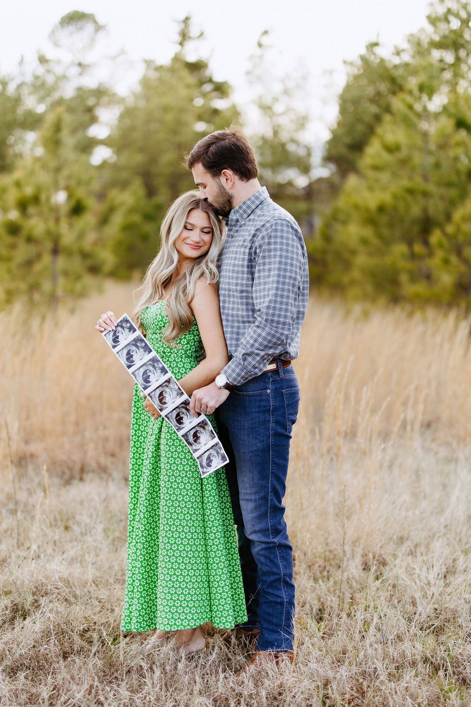 Couple stands together in golden field holding letter banner, with woman wearing polka dot green dress.
