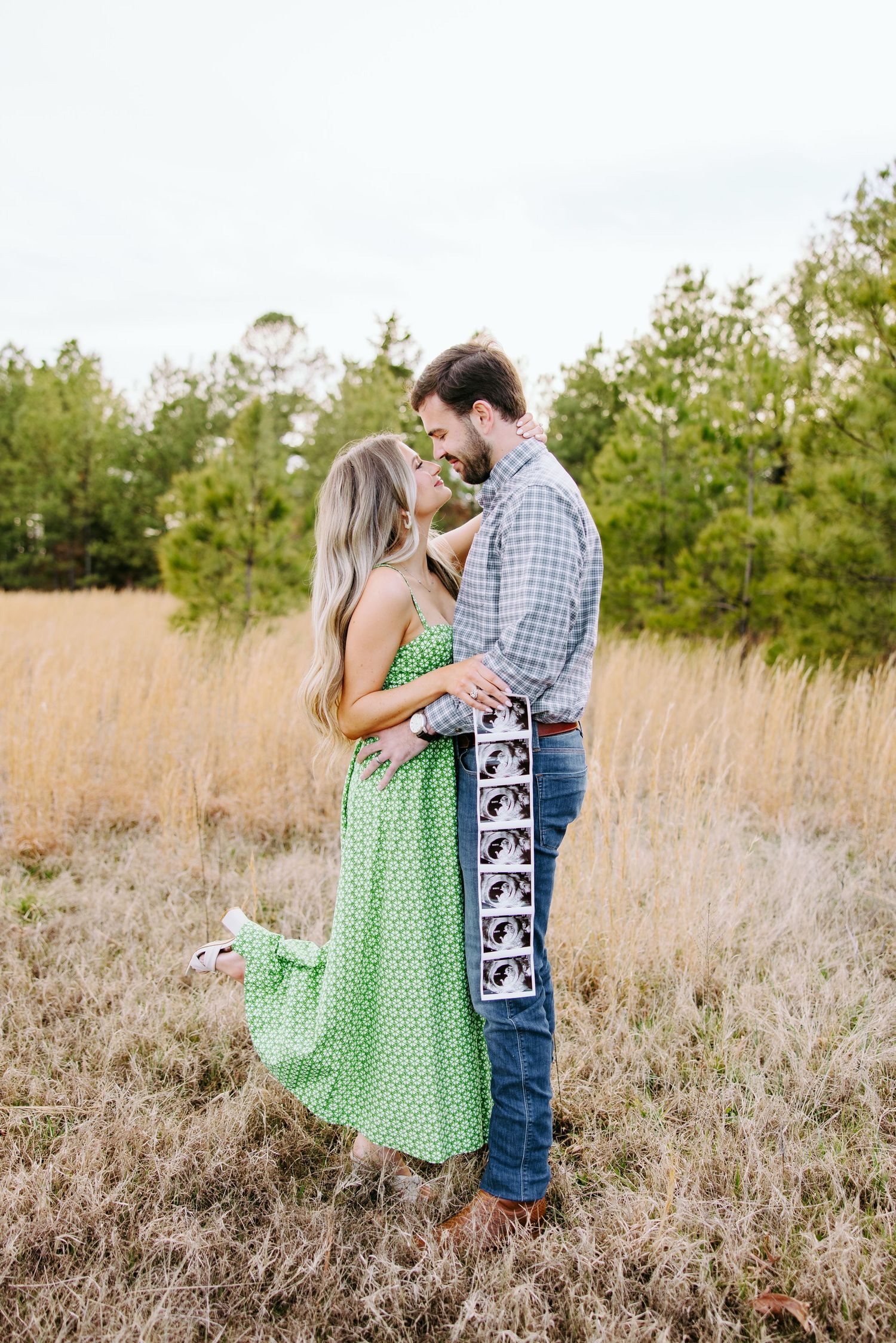Romantic couple embraces in golden field with trees, woman wearing green polka dot dress and man in plaid shirt.