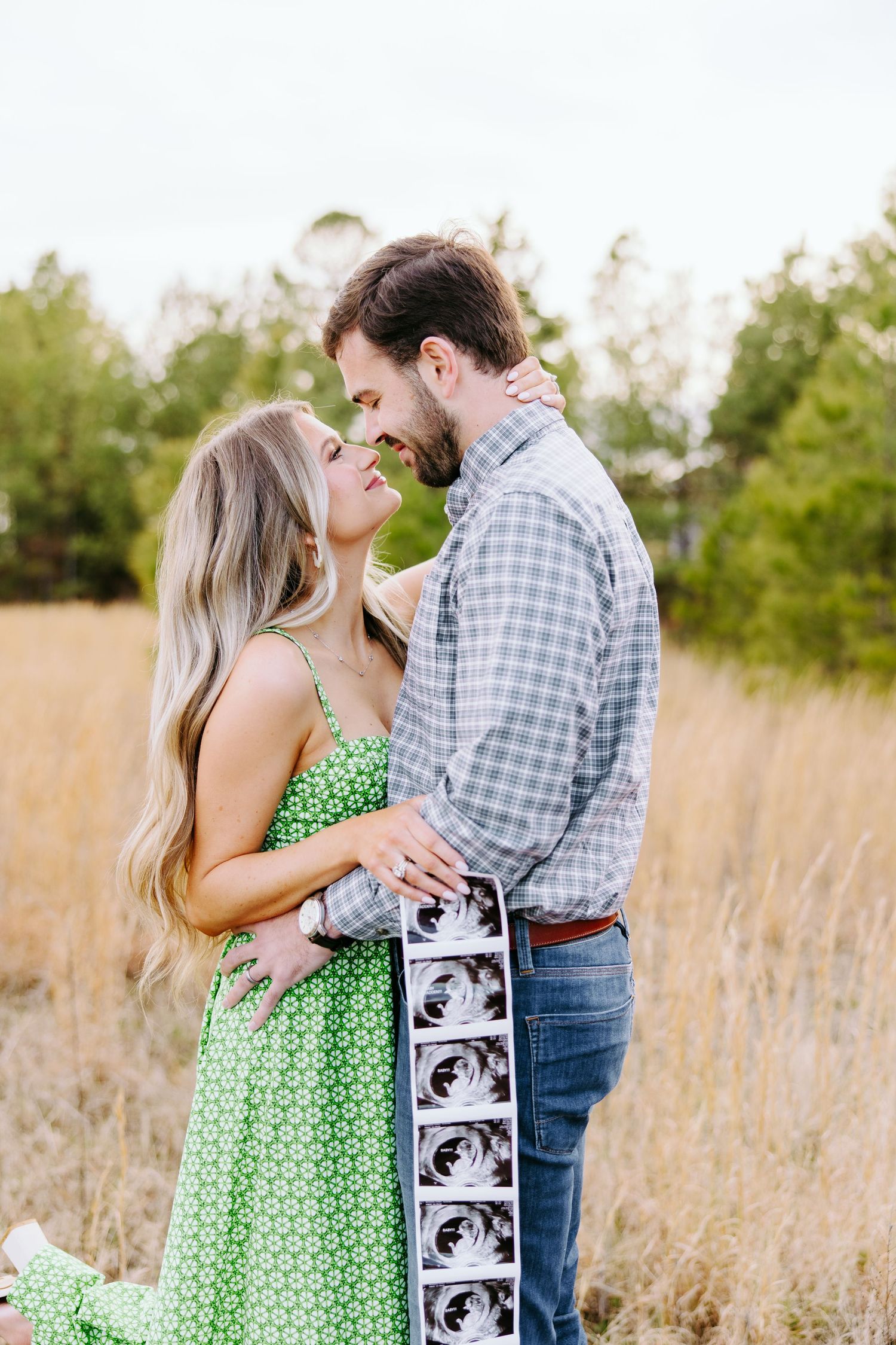 A couple shares an intimate moment in a grassy field while holding ultrasound photos.
