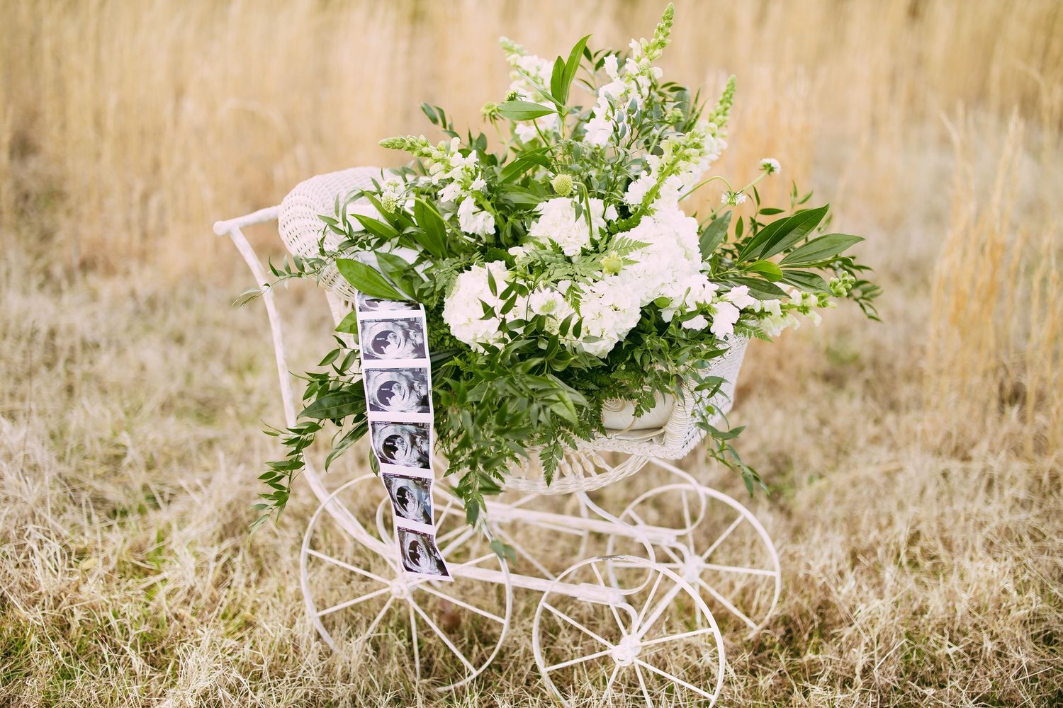 A decorative white bicycle planter filled with lush white flowers and greenery sits in a field of golden grass.