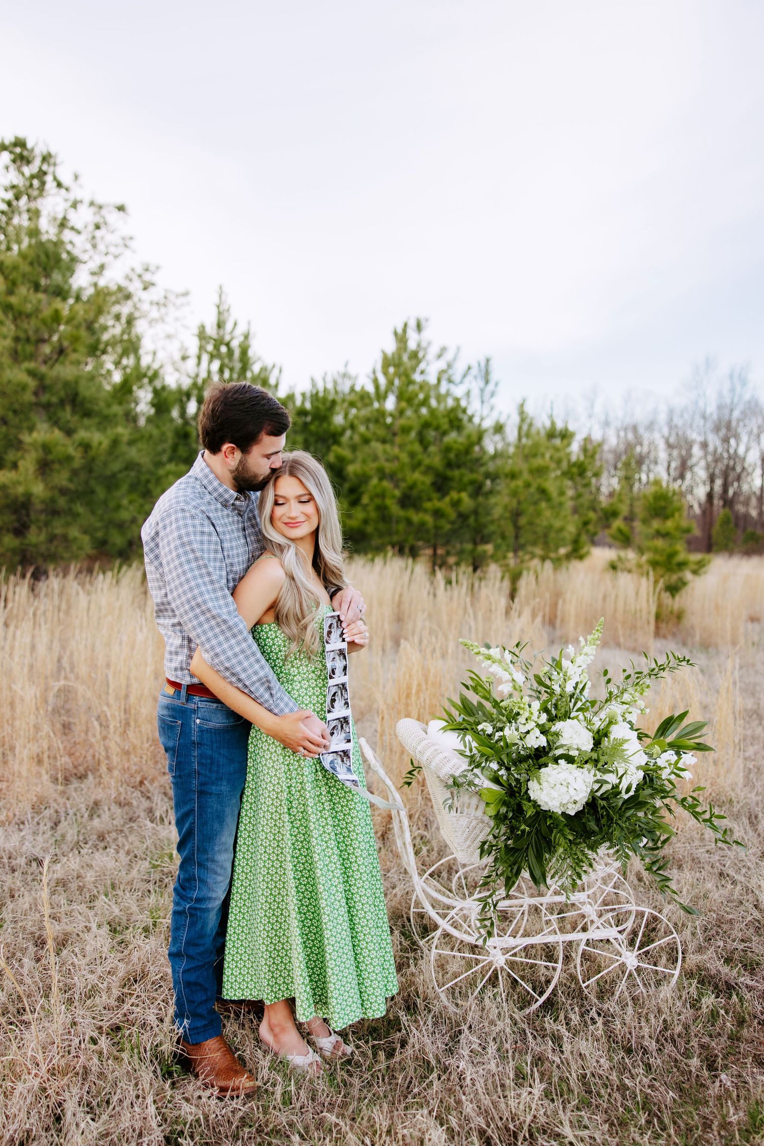 A couple embraces in a rustic field setting with dried grass and a beautiful white floral arrangement nearby.