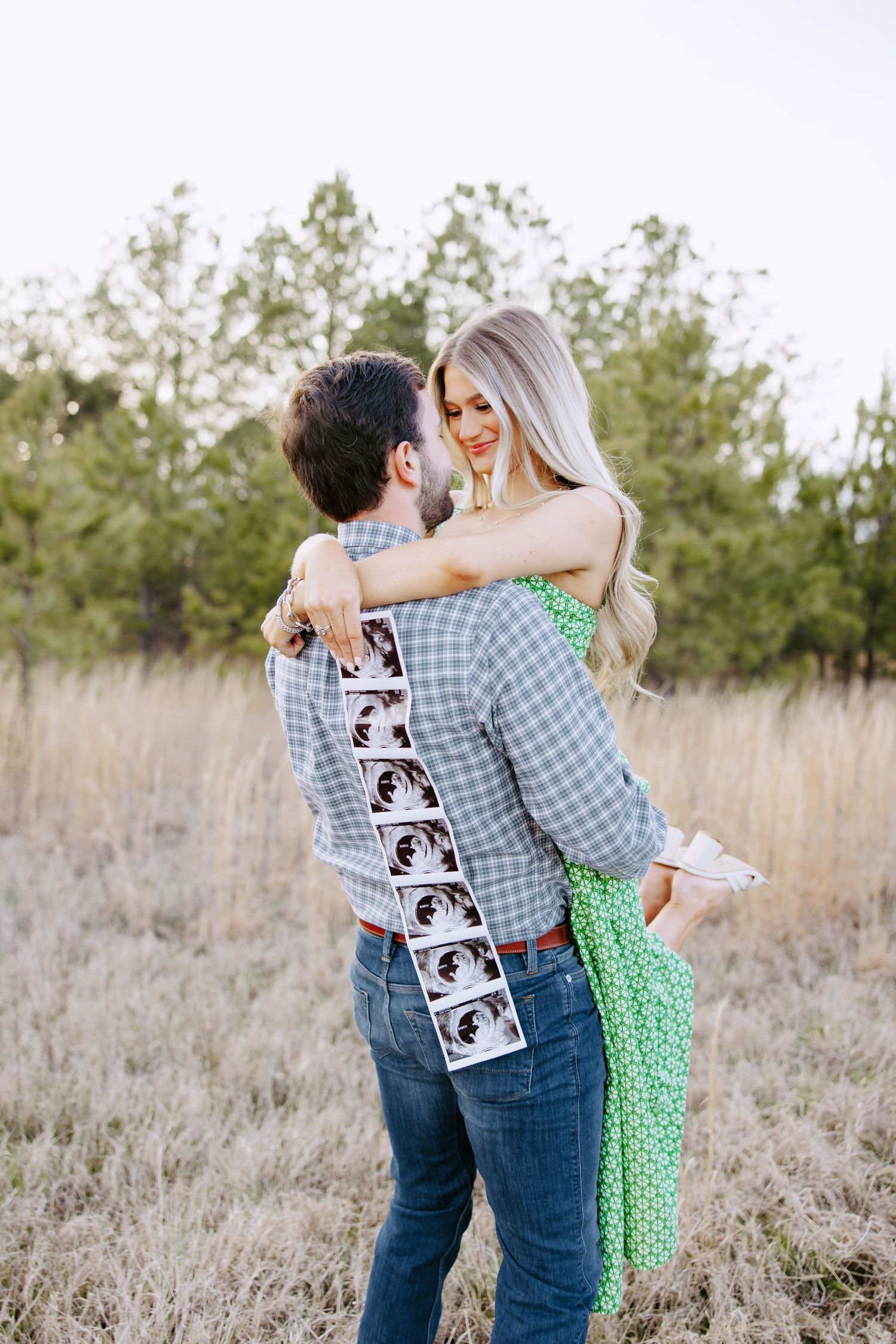A couple embraces in a field of tall grass while wearing casual attire with ultrasound photos displayed on the back.