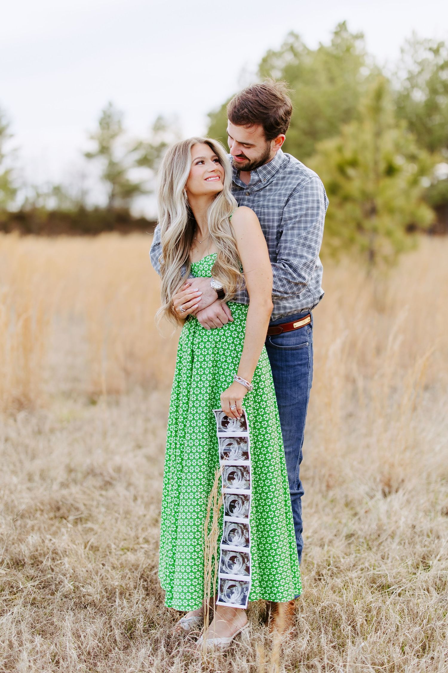 A couple stands together in a golden field while holding ultrasound photos on a sunny afternoon.