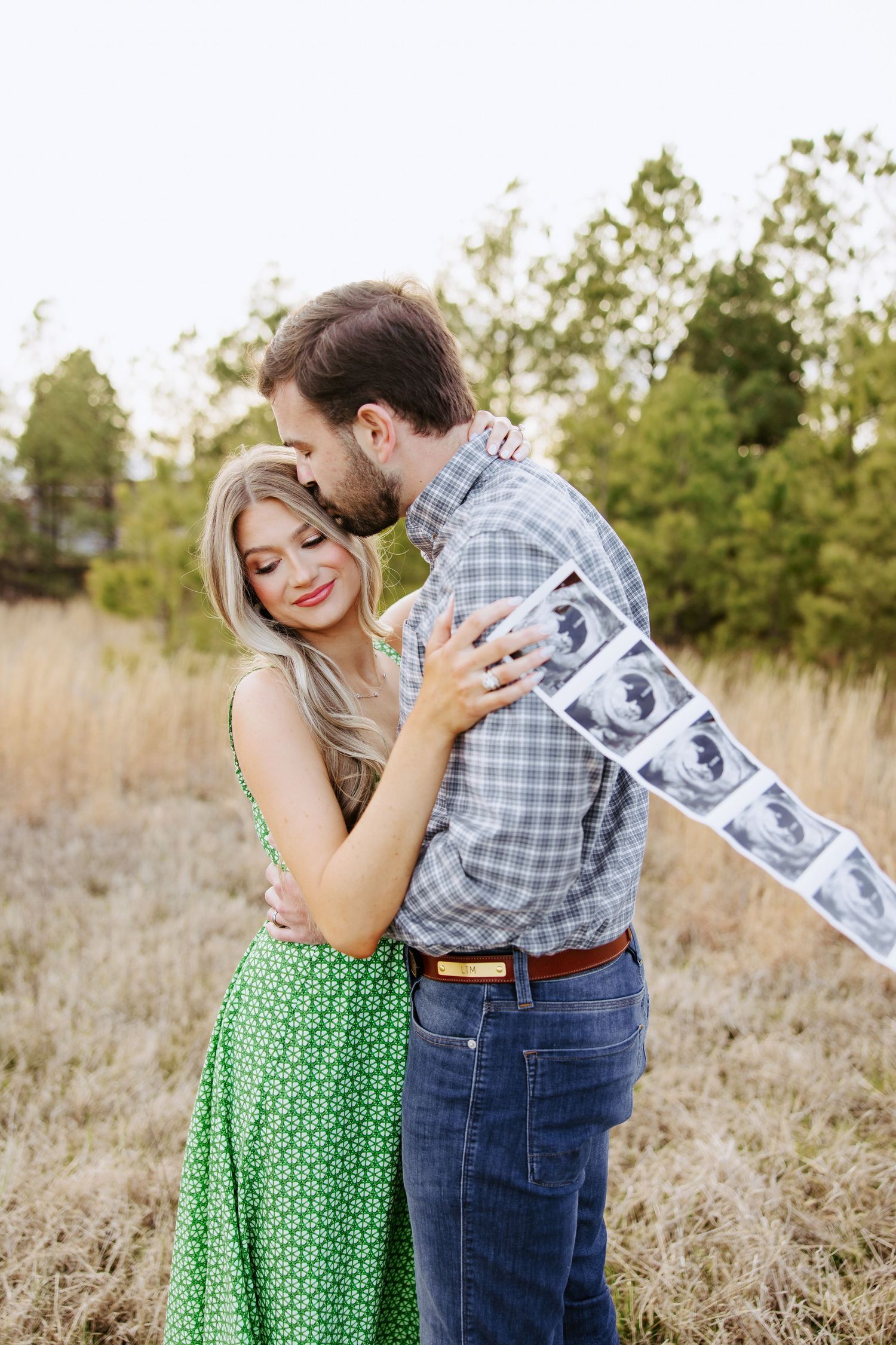 Couple embraces in a field while holding ultrasound photos in a sweet pregnancy announcement photo.