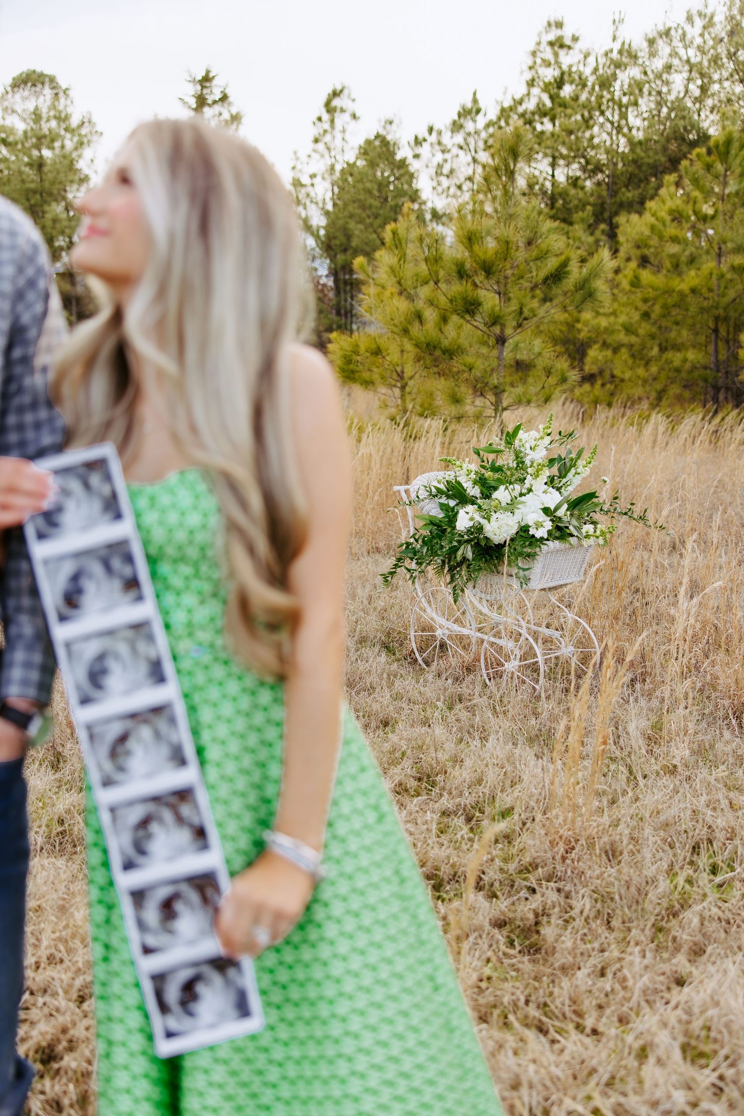 Person in green polka dot dress holds ultrasound photos in tall grass field with floral bouquet arrangement nearby.