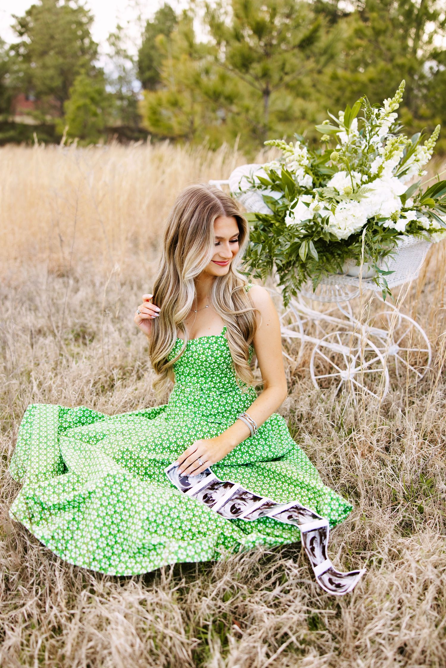 A person in a green polka dot dress sits in a field of tall grass next to white floral arrangements.