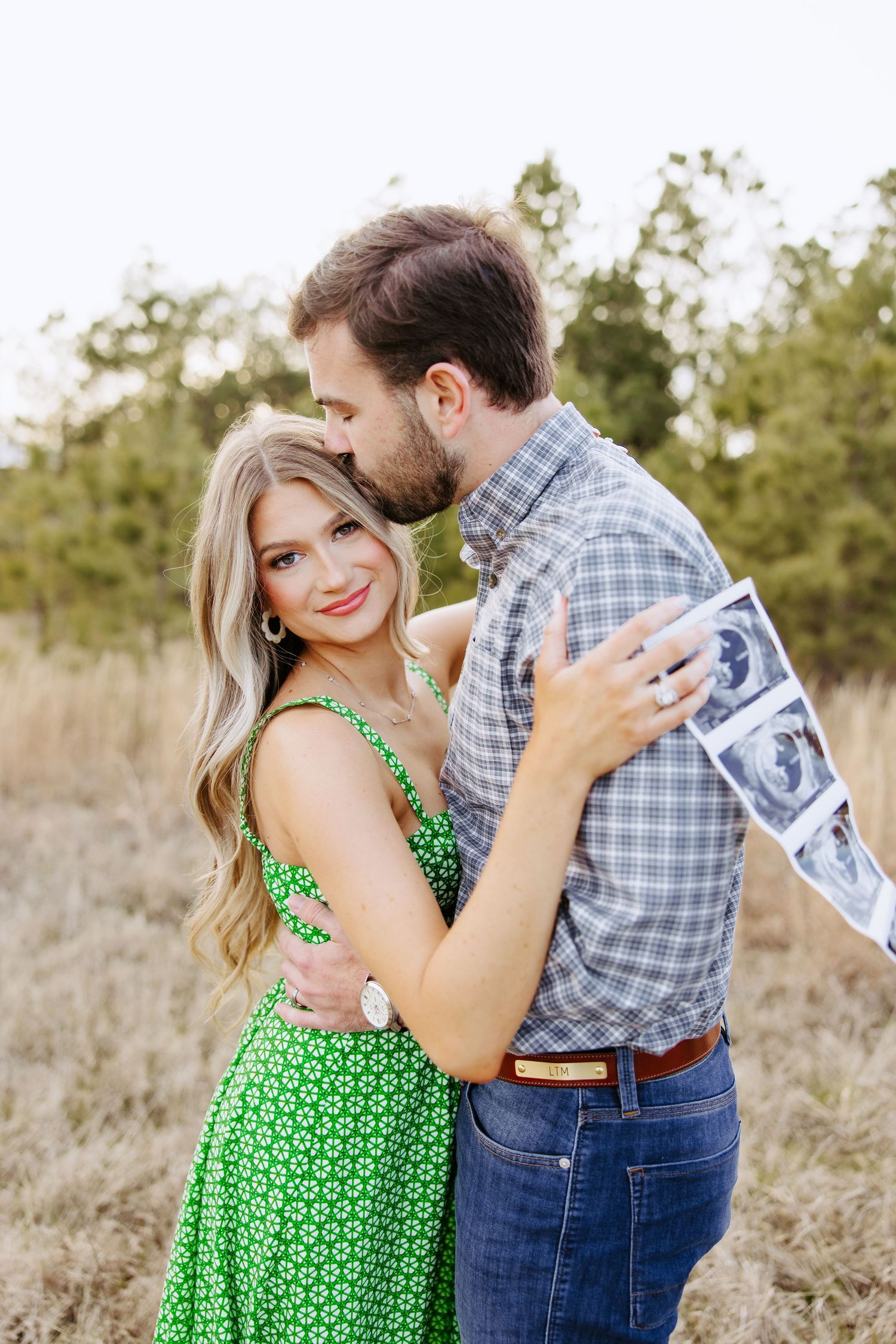 A couple in a field embraces while holding an ultrasound photo, wearing a green polka dot dress and plaid shirt.