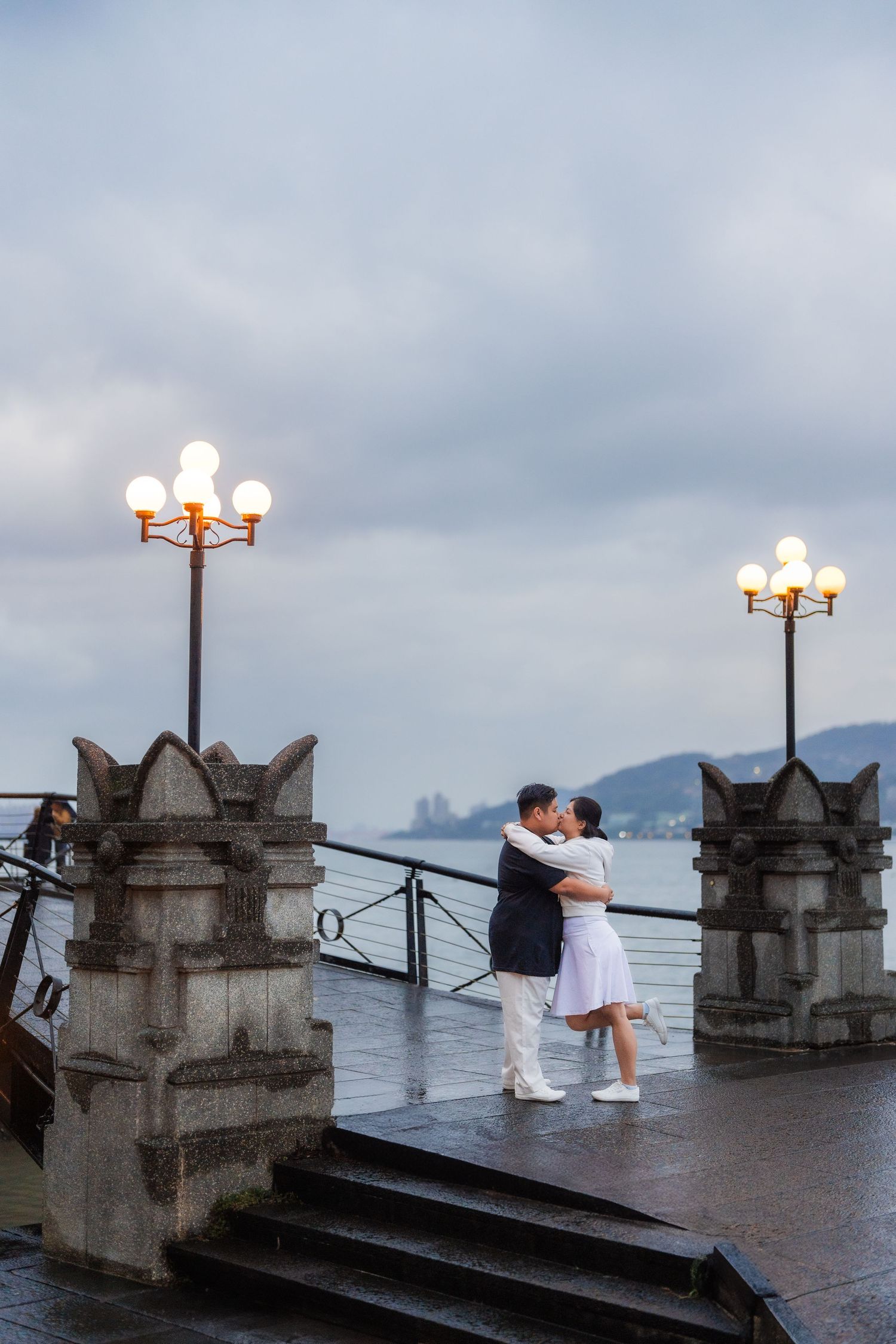 A couple embraces on a romantic stone terrace with ornate lamp posts and cloudy mountain views in the background.