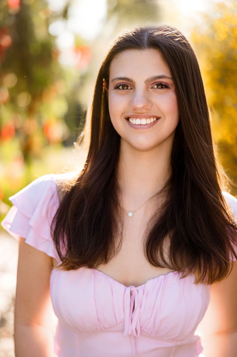 A portrait photo of a smiling person with long dark hair wearing a pink dress in front of a warm autumn background.