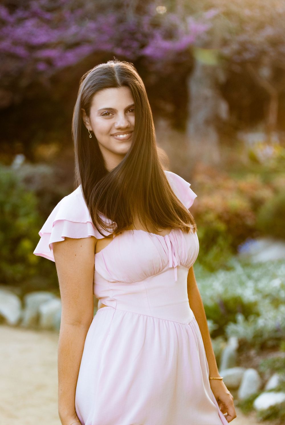 A person in a light pink off-shoulder dress poses outdoors against a colorful garden backdrop during golden hour.