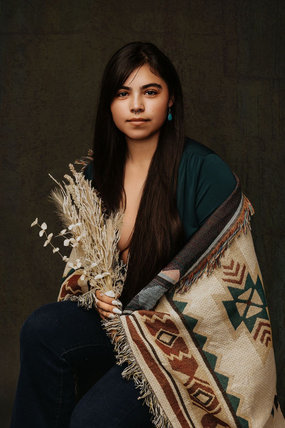 A person wearing a teal top and traditional Native American blanket holds dried pampas grass in a studio portrait.