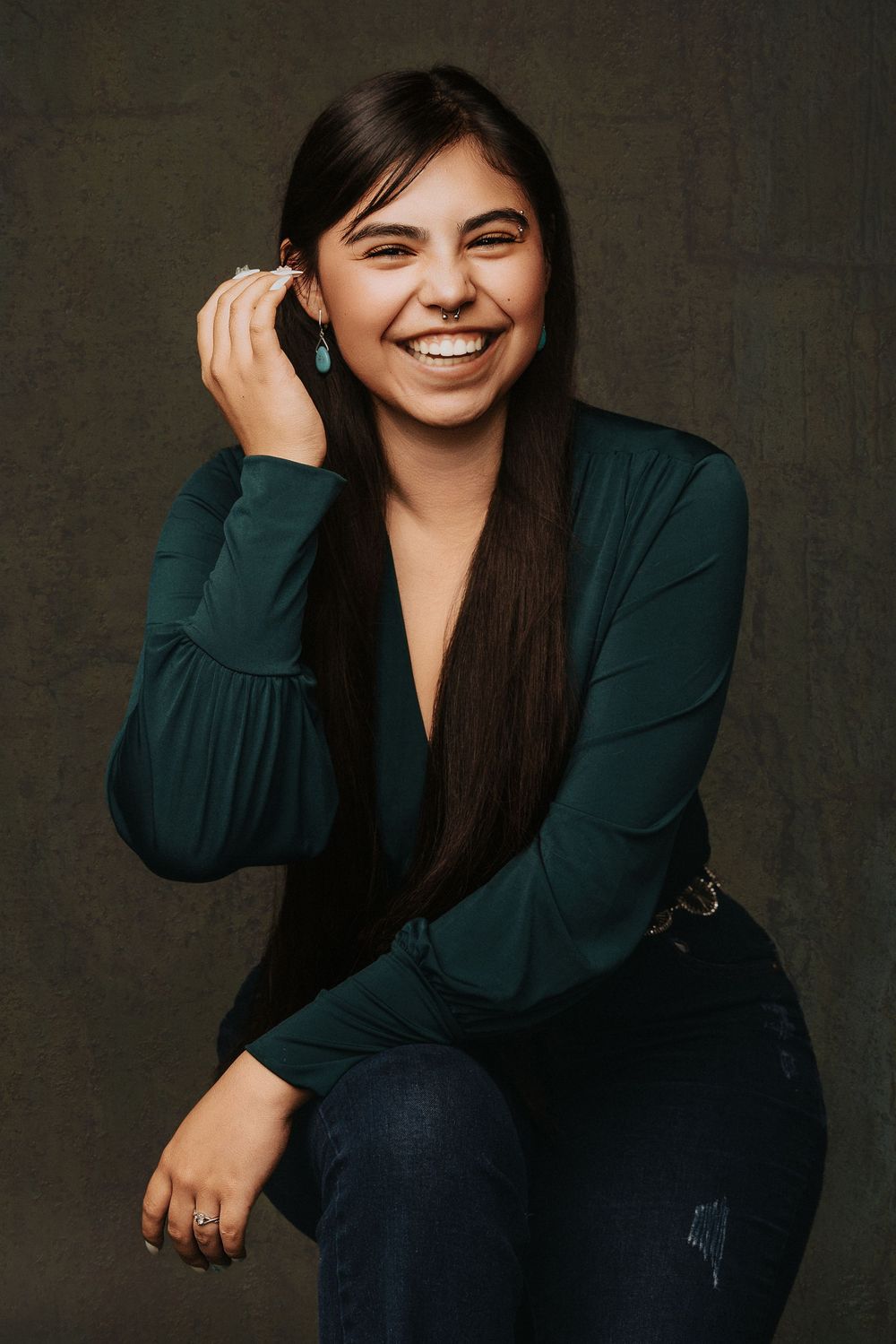 Professional portrait of a smiling person with long dark hair wearing a deep green top against a dark background.