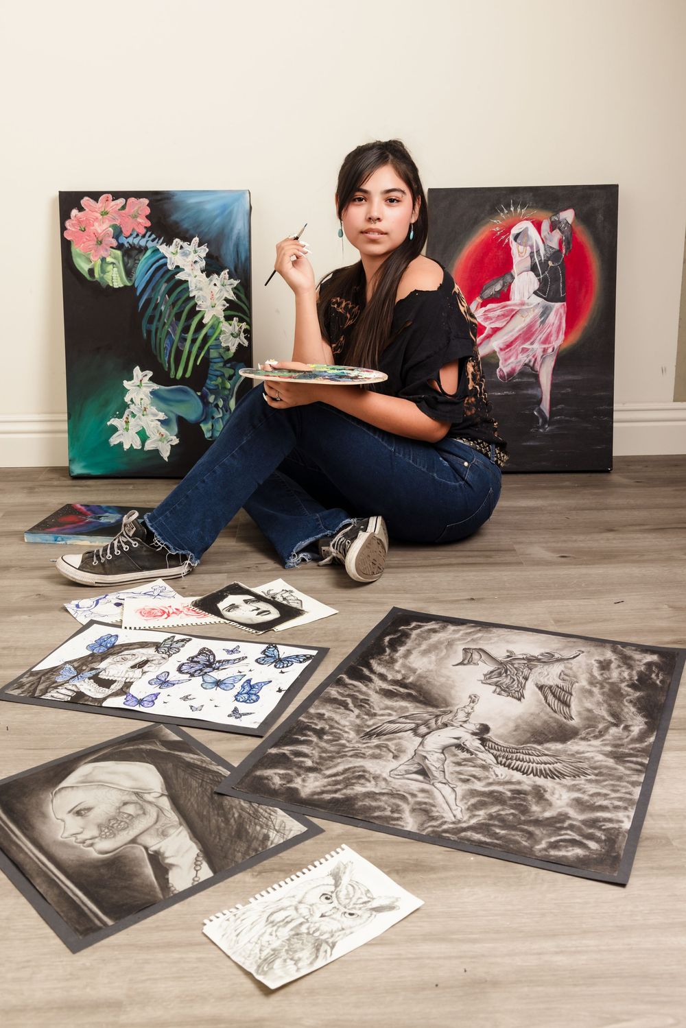 Artist sits on floor surrounded by various artworks including floral paintings and sketches while holding a sketchbook.