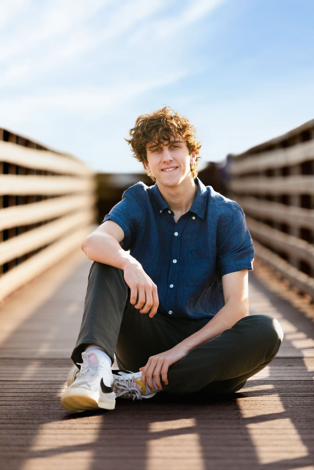 A person in a blue button-up shirt sits cross-legged on a wooden boardwalk while sunlight casts dramatic shadows.