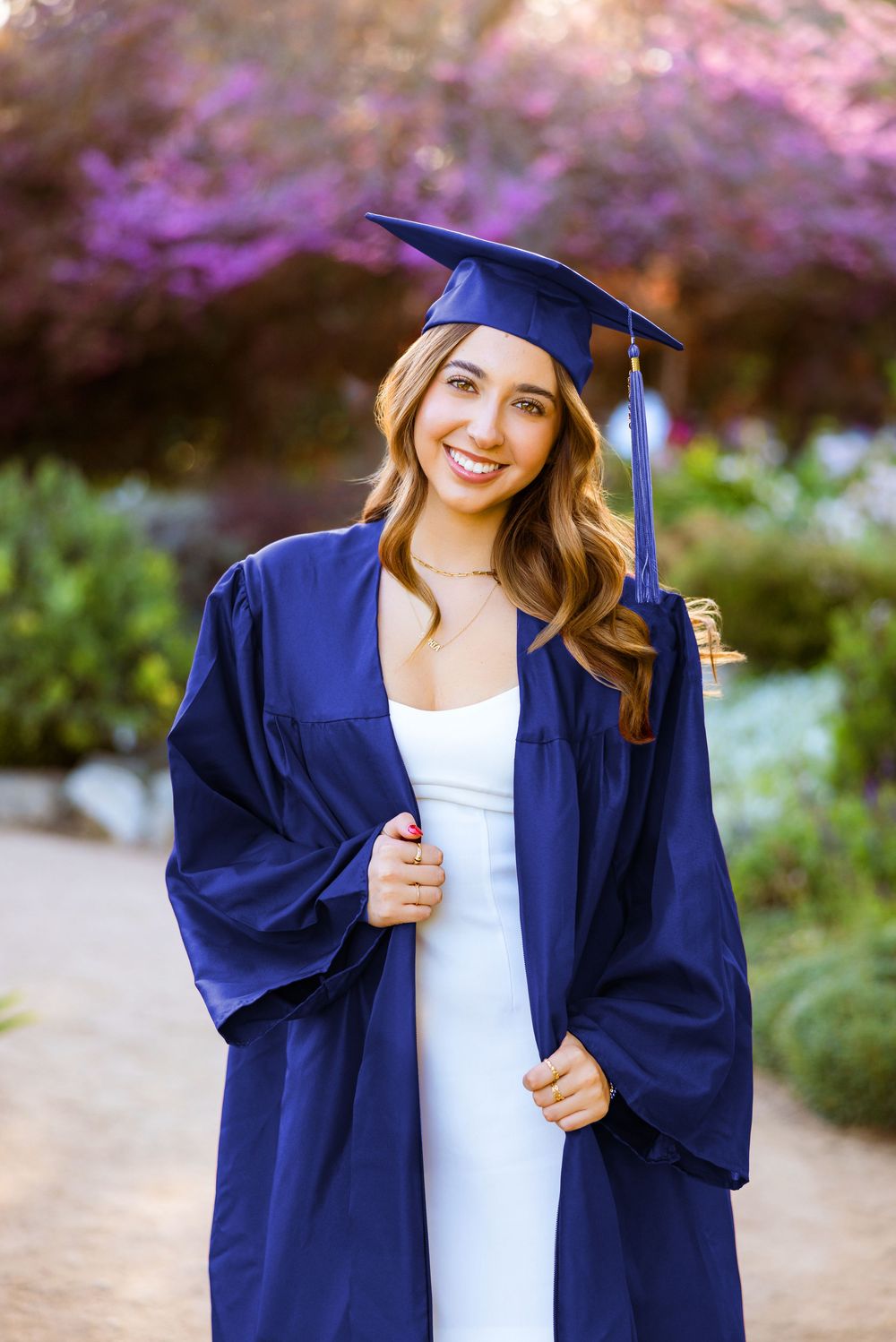 A graduate wearing blue cap and gown stands outdoors with blooming pink flowers in the background.
