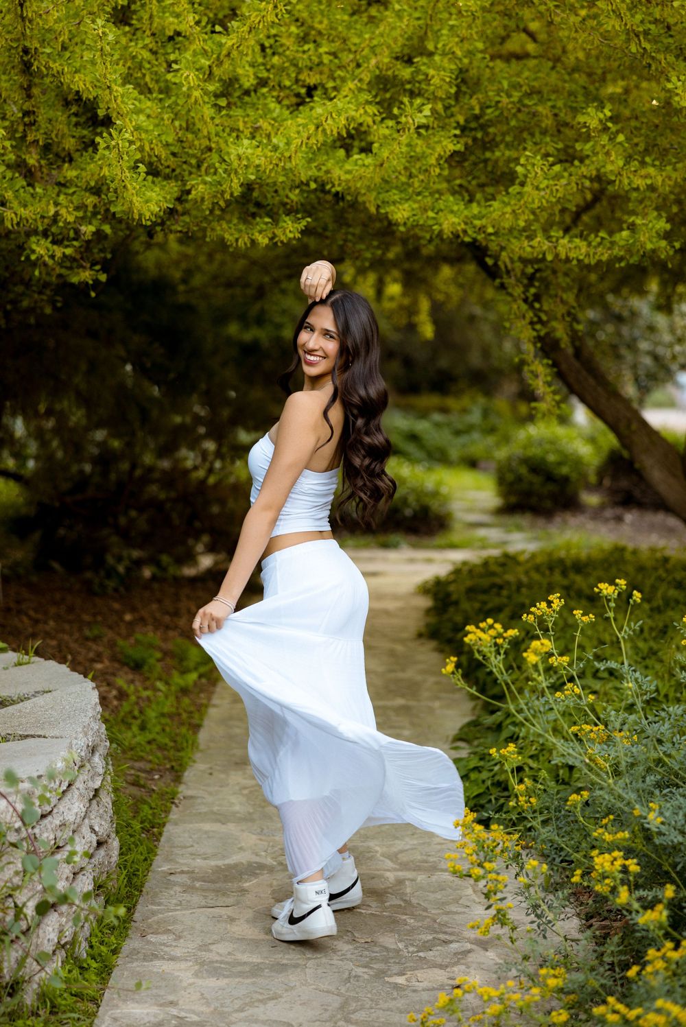 Woman in flowing white outfit poses gracefully on a garden path surrounded by lush greenery and yellow flowers.