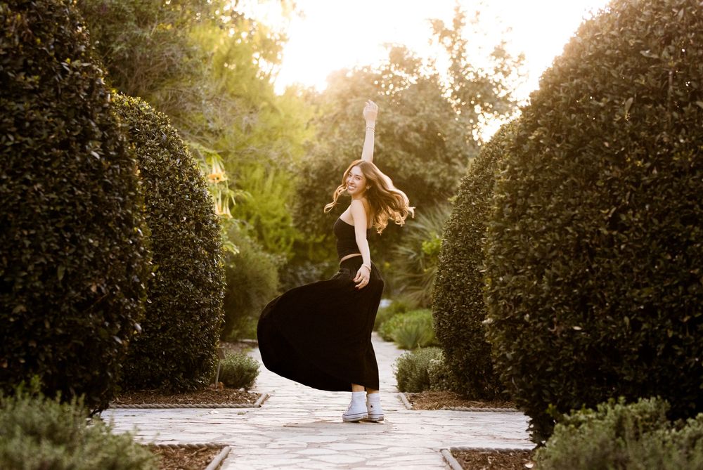 A dancer in a black dress twirls gracefully on a garden path surrounded by manicured hedges at golden hour.