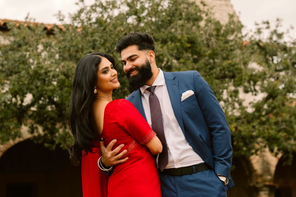 A couple in formal attire, with the woman in a red dress and man in navy suit, embrace outdoors against greenery.