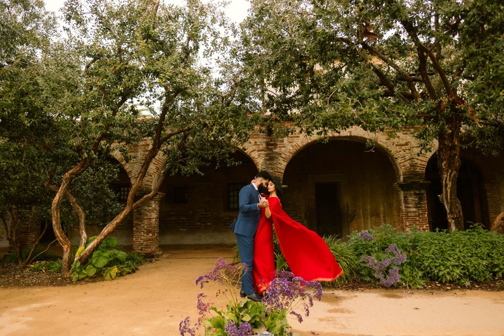 Romantic couple embraces at golden hour under stone archway with red dress flowing among garden blooms and trees.