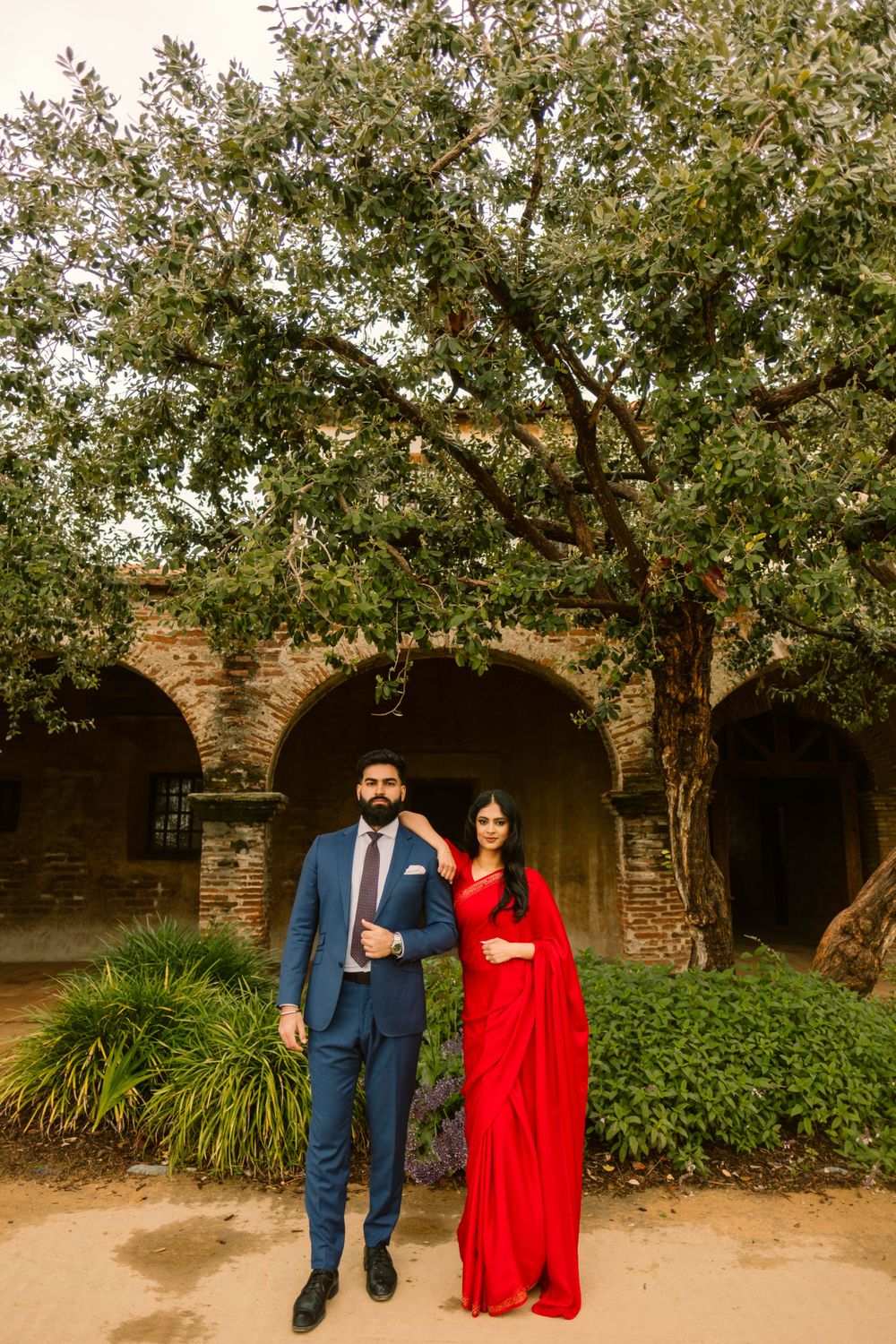 A couple in formal attire standing in front of stone archways surrounded by lush greenery and trees.