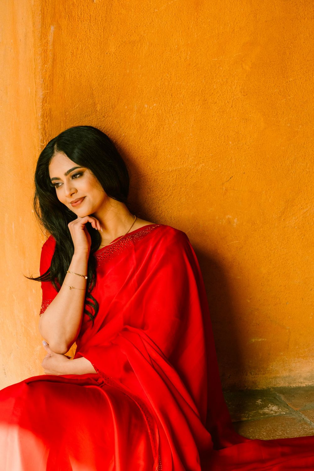 A person in a vibrant red dress poses thoughtfully against a warm orange wall, creating a striking color contrast.