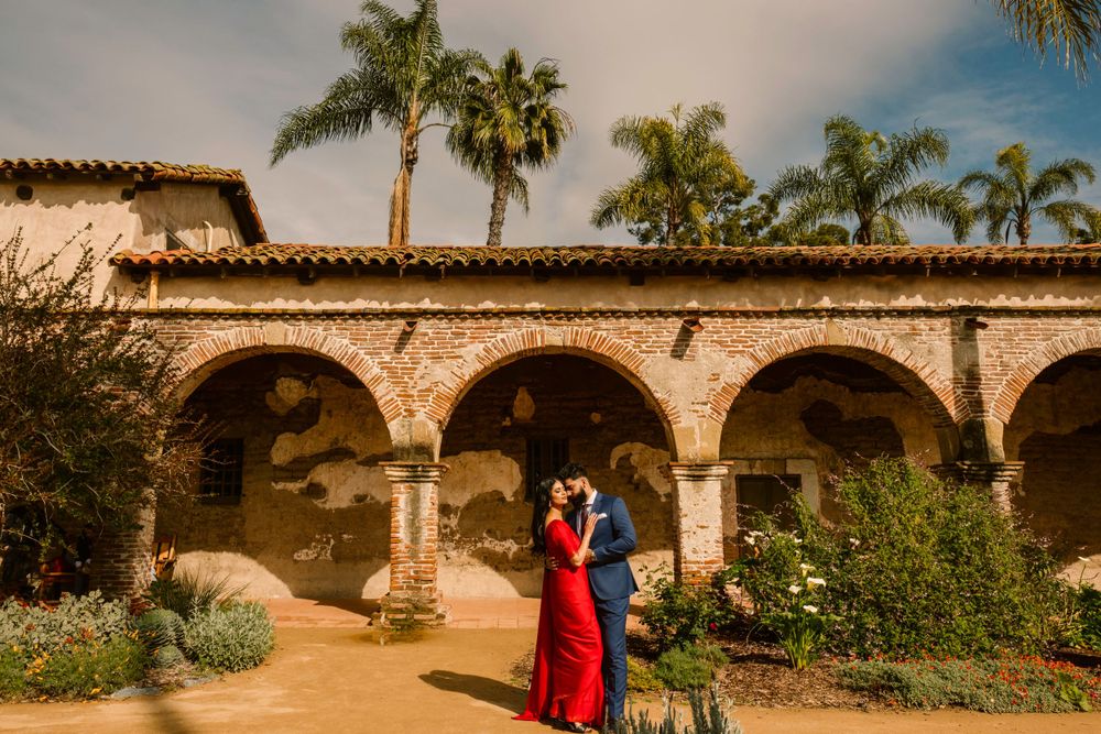 Spanish colonial arches frame couple in red dress beneath swaying palm trees at sunset mission courtyard.
