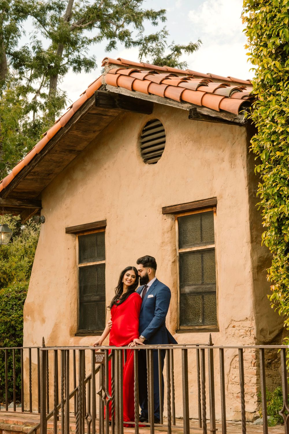 A couple stands on a balcony outside a Spanish-style stucco building with terracotta roof tiles and dark trimmed windows.