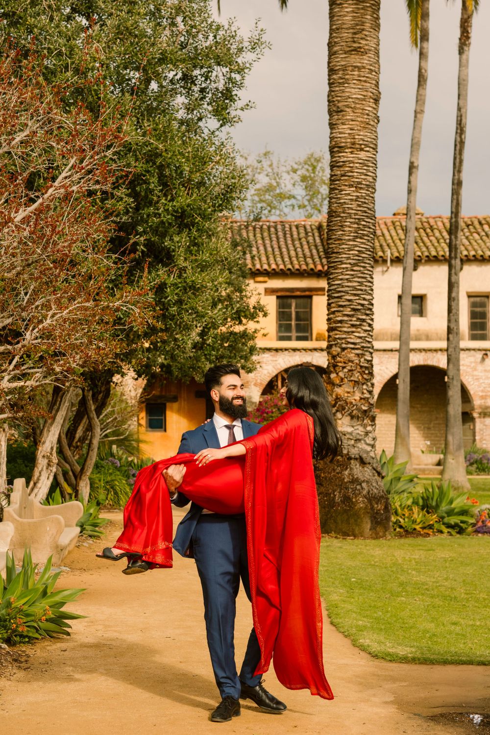 A romantic moment at a Spanish colonial style courtyard with palm trees and red flowing gown against beige architecture.