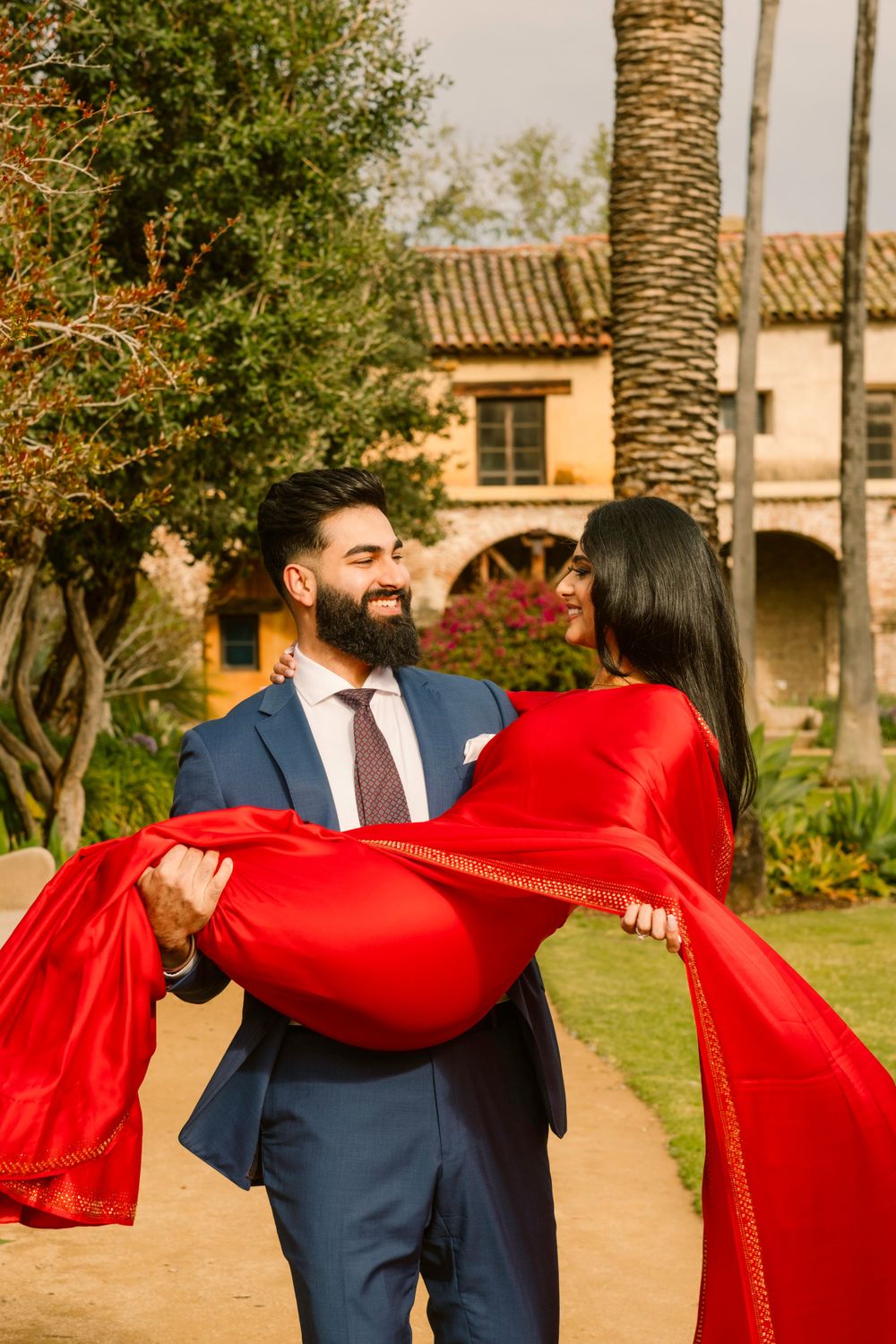 A couple shares a romantic moment in a garden with palm trees, as person in blue suit carries person in flowing red dress.