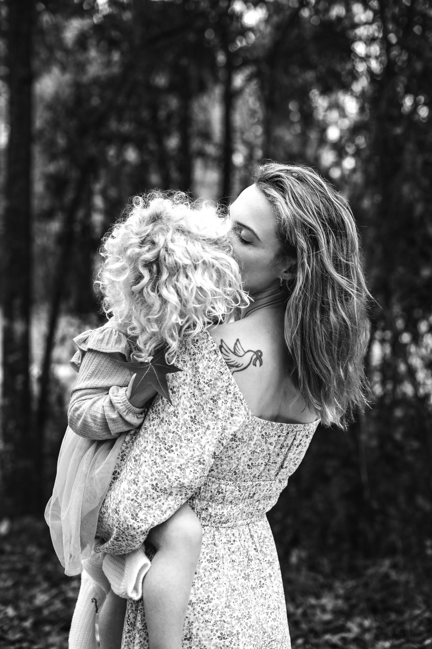 A tender black and white portrait of a mother in a floral dress embracing her curly-haired child in a forest setting.