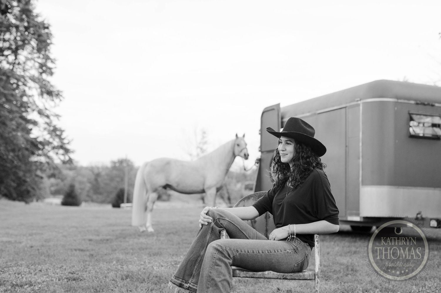 Black and white portrait showing a peaceful rural scene with a horse trailer and white horse in the background.