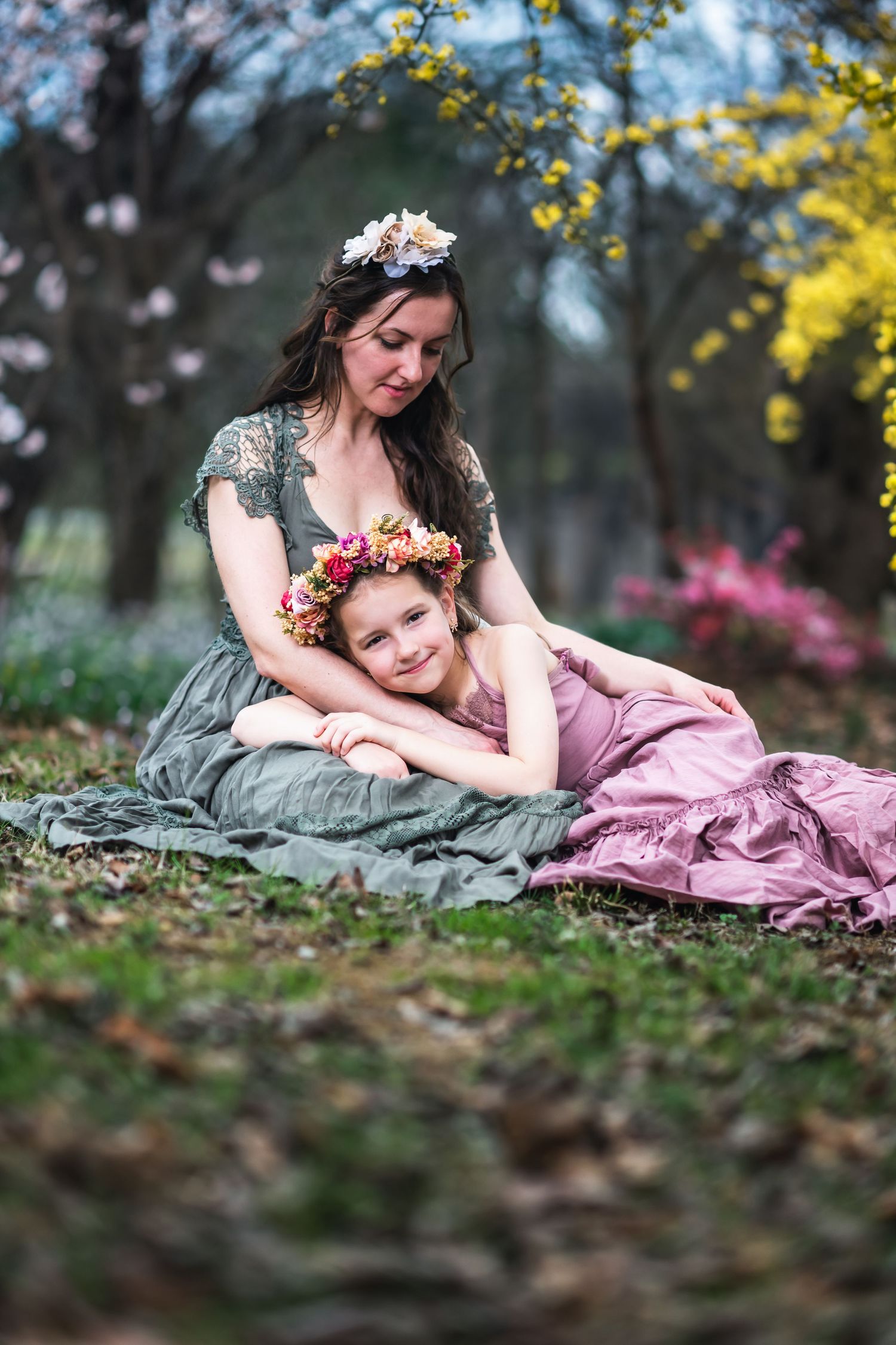 Two women in vintage dresses and flower crowns sit together in an ethereal spring garden setting with blooming flowers.