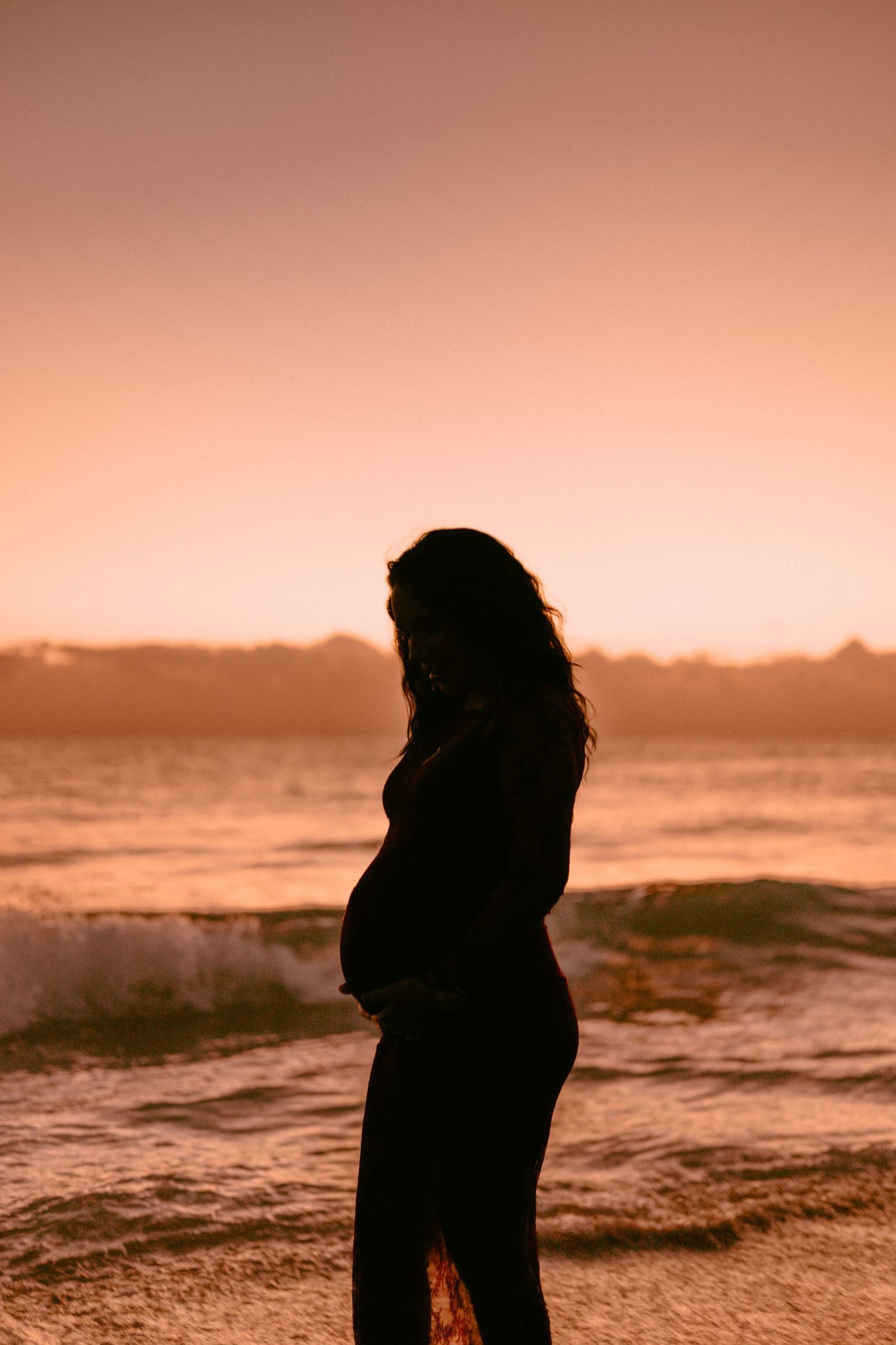 Golden image of a pregnant women posing in front of a sunrise on the beach shore with waves crashing in the background.