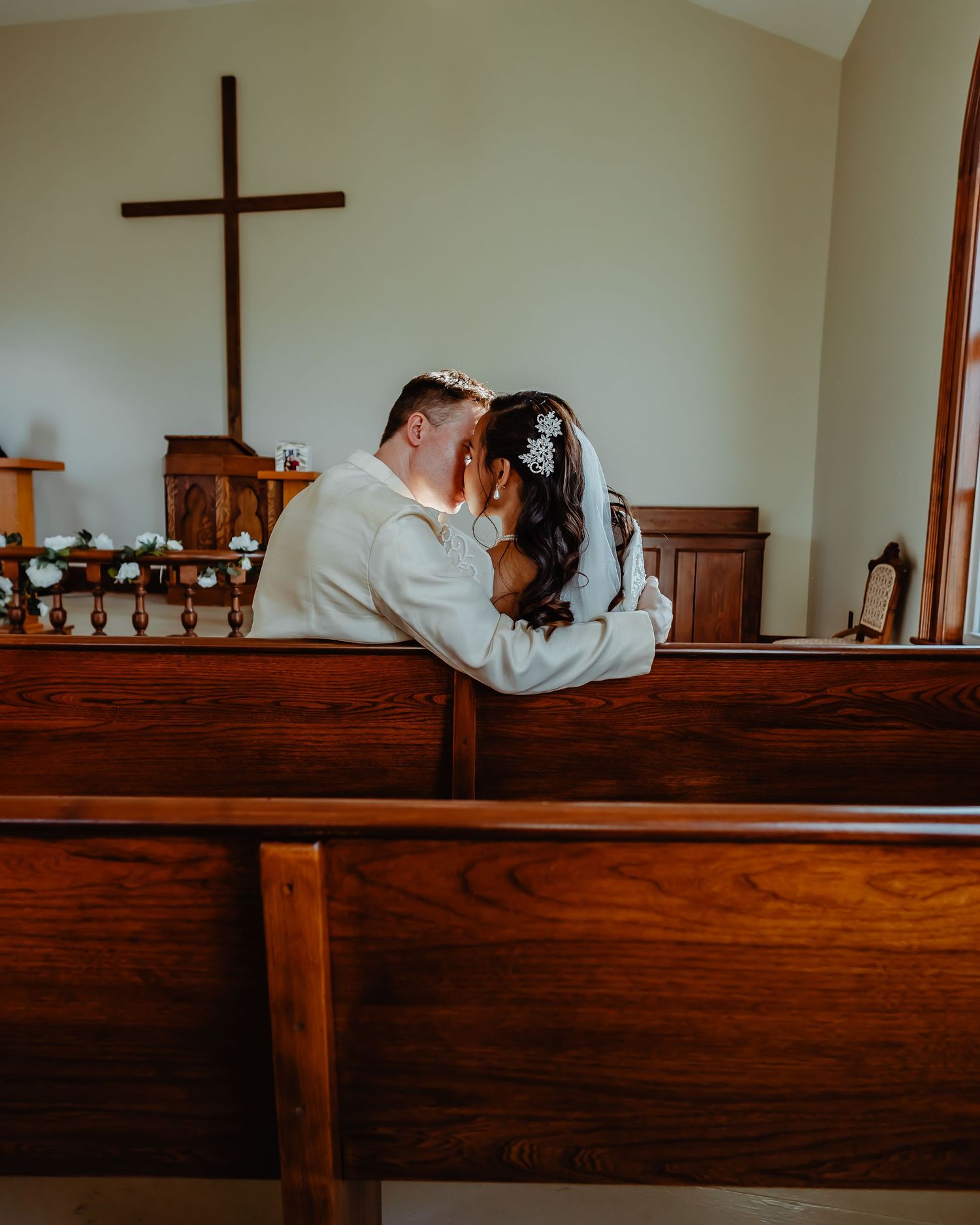 A couple shares an intimate moment while sitting in a wooden church pew beneath a wooden cross on the wall.