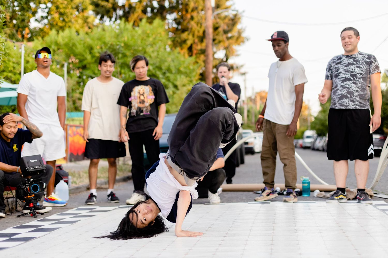 A street break dancer performs a dynamic move on pavement while onlookers watch in an outdoor setting.