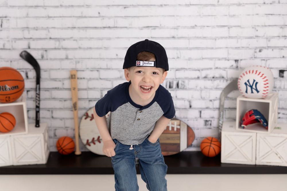A young sports fan poses energetically in front of a white brick wall with basketballs and a Yankees baseball on display.
