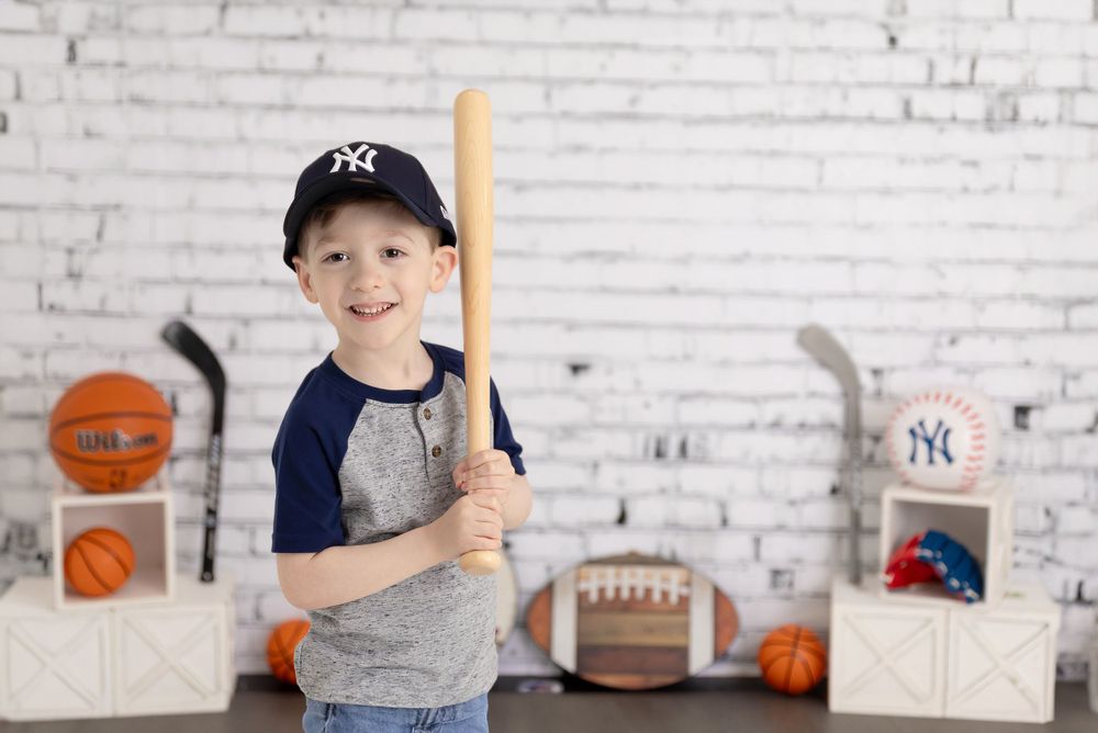 Young baseball fan in NY Yankees cap stands with bat against white brick wall with sports equipment display.