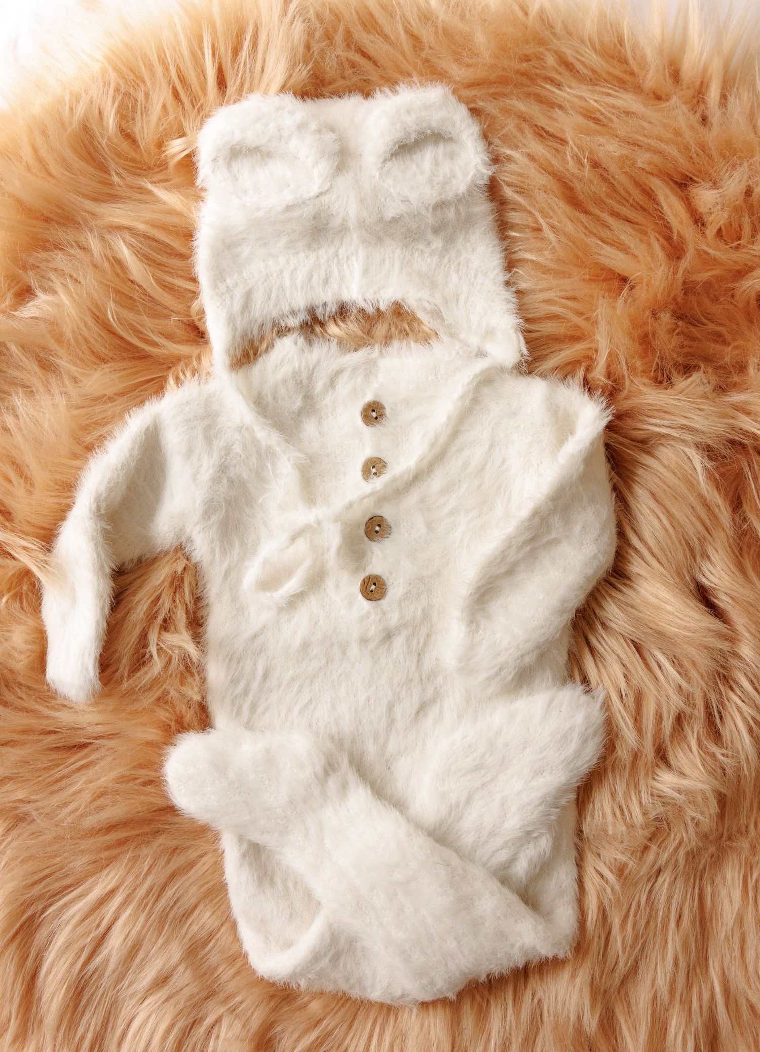 White plush baby snowsuit with ears on hood and wooden buttons laid out on soft peach-colored fur background.