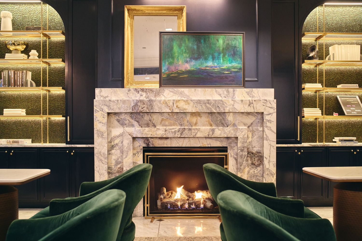 Modern luxury lounge with marble fireplace, emerald velvet chairs, and built-in shelving displaying books and decor.