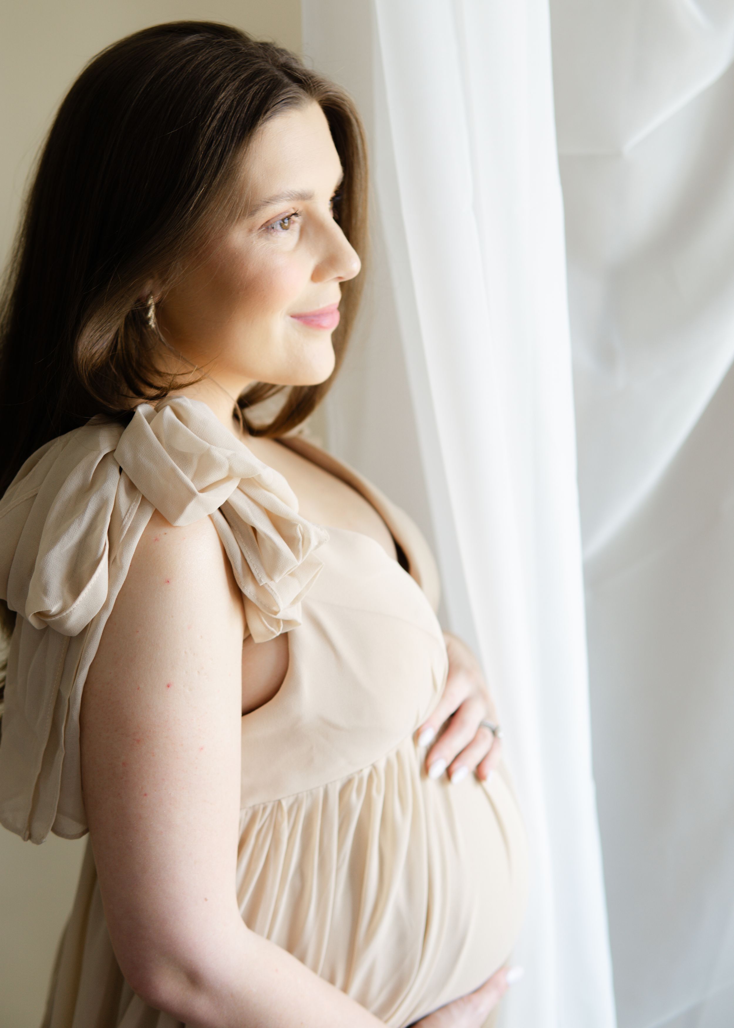 Tender maternity portrait, expecting mother framed by window light, wearing flowing dress with bow accent.