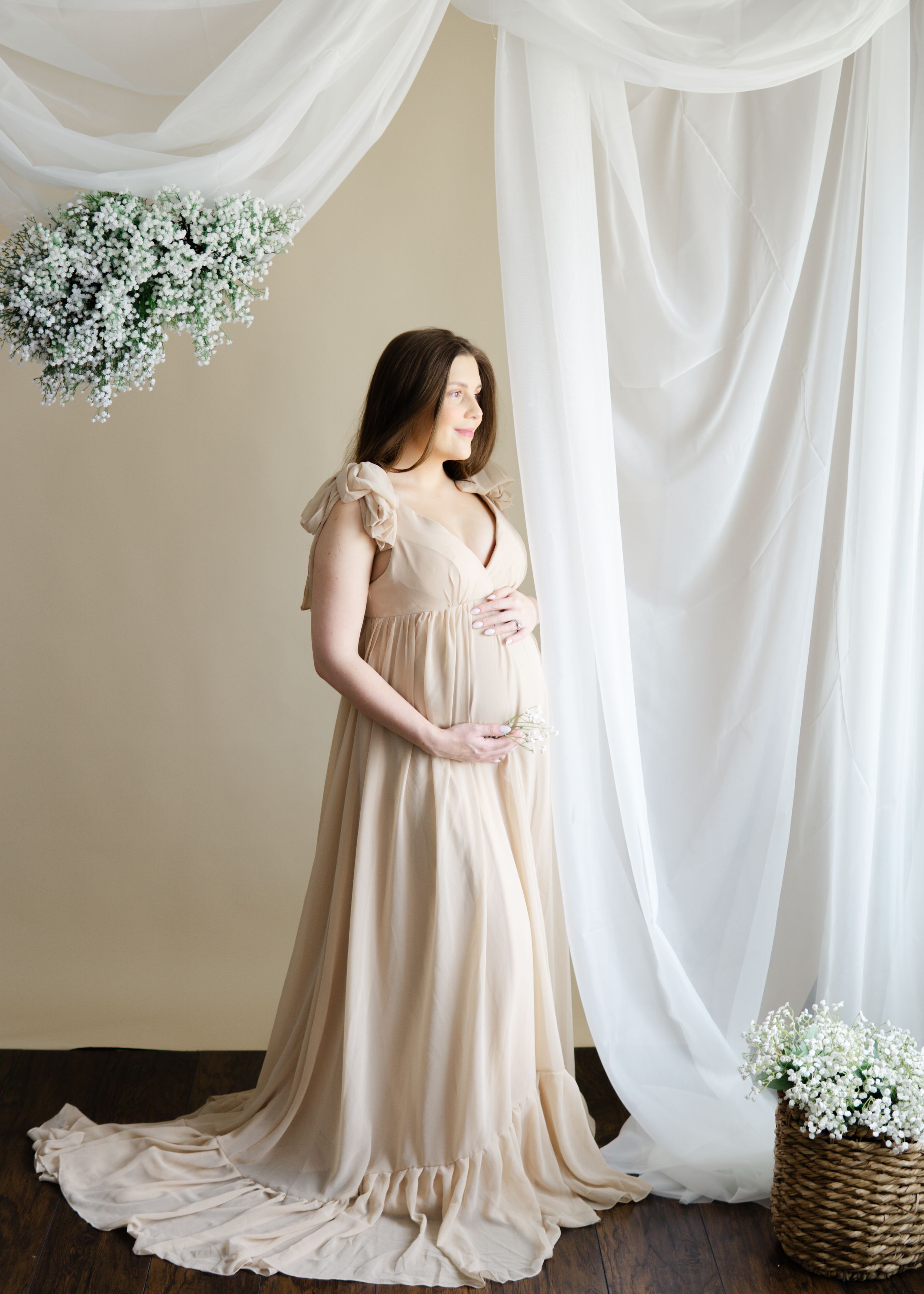 Full-length maternity portrait, expecting mother in flowing beige gown, white draped backdrop with floral accents