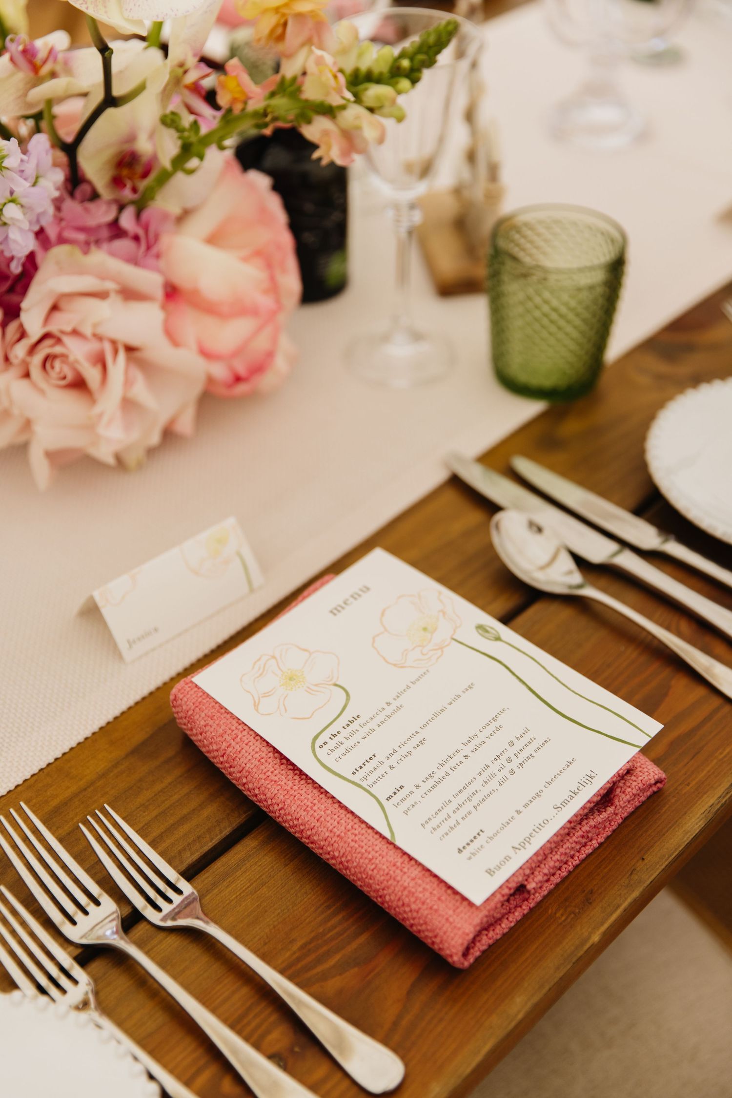Elegant table setting with pink rose centerpiece, menu card with coral napkin, and vintage silverware on wooden surface.