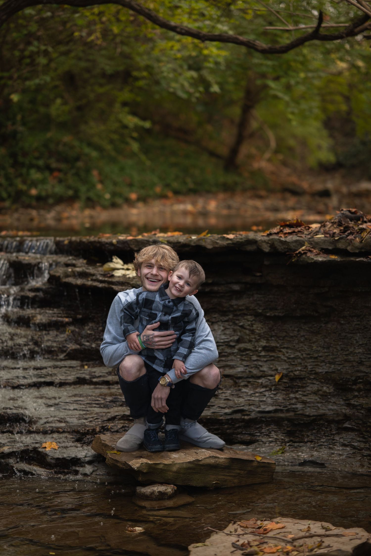 Two people sit together on dark rocks in a stream surrounded by green foliage in a forest setting.