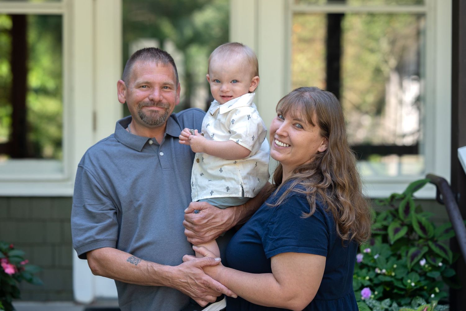 A family smiles while posing together outside their home with green plants and flowers in the background.