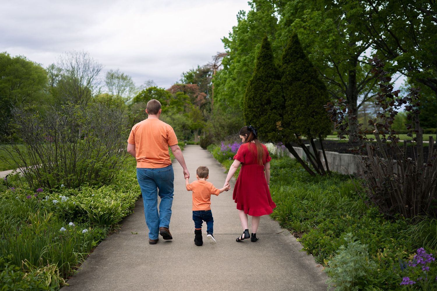 Family of three walking hand-in-hand down a garden path surrounded by lush green trees and plants.