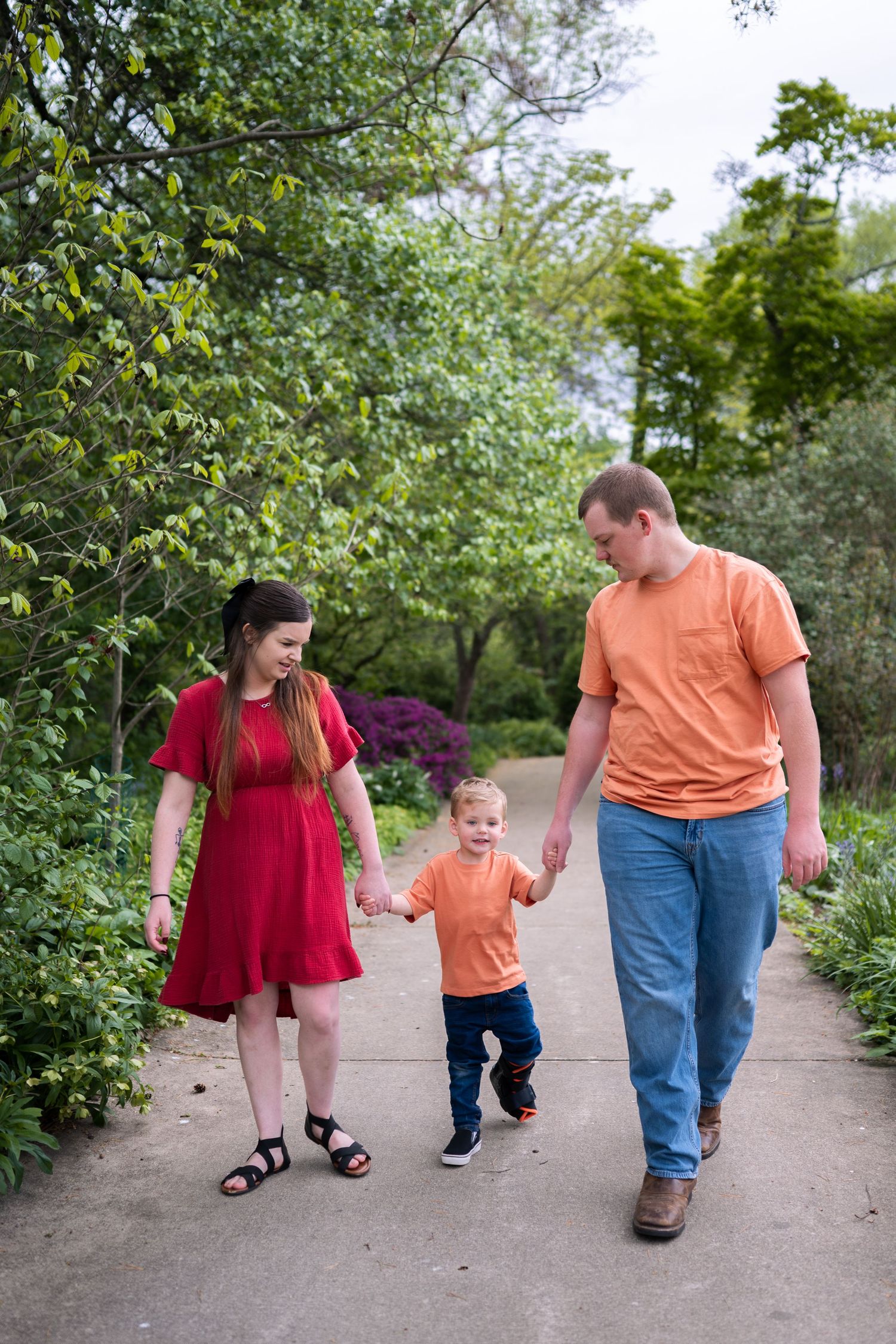 Family of three walking hand-in-hand on a garden path, parents in red and orange shirts with toddler between them.