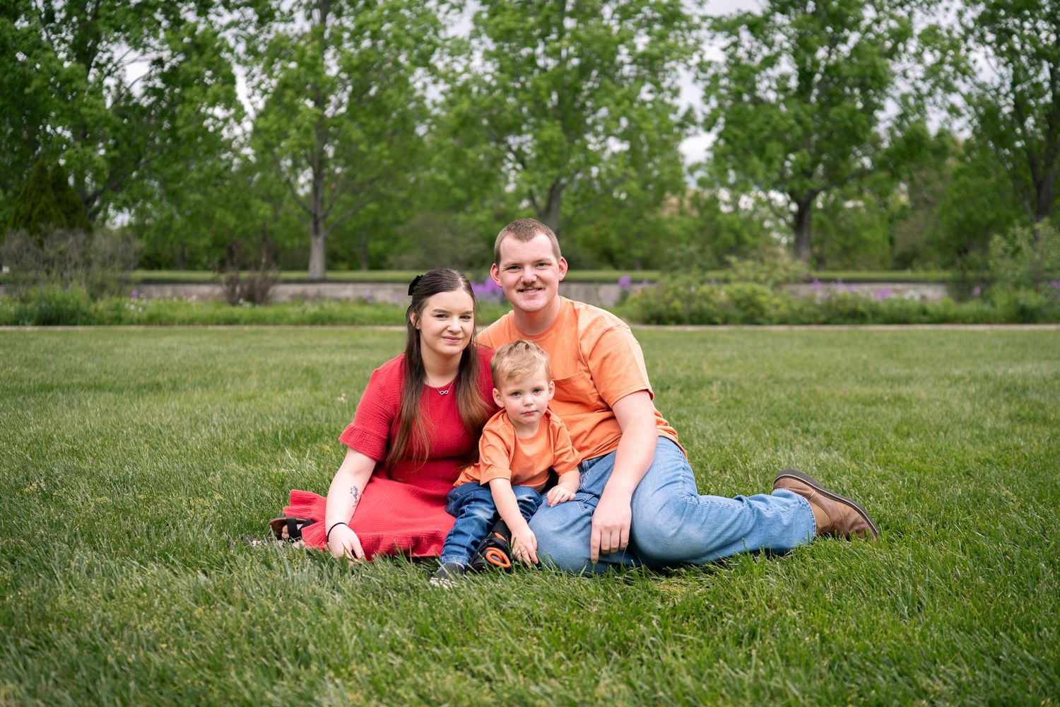 Family of three sitting on green grass in a park, smiling couple with young toddler boy on a sunny day.