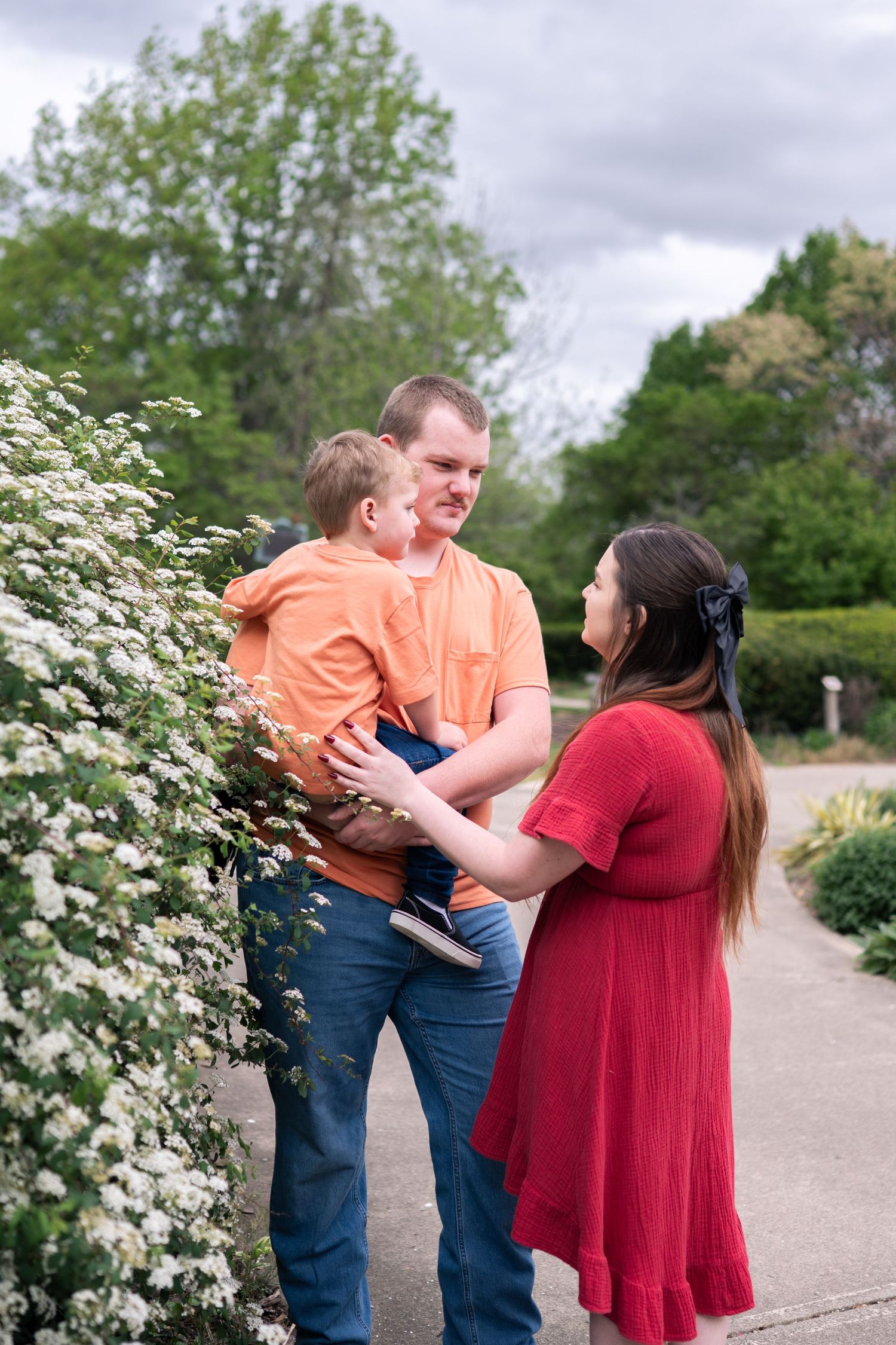 Family of three enjoying a spring day outdoors, father holding toddler in orange shirt, mother in red dress.